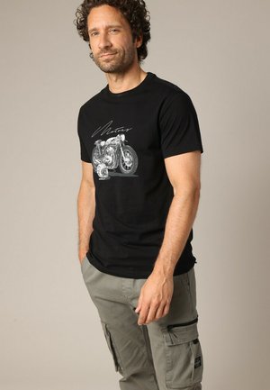 Zwarte t-shirt met een grafisch ontwerp van een motorfiets, met witte tekst die "Motor" zegt, gecombineerd met lichtgroene cargobroeken.