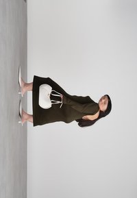 Donna in un abito marrone scuro con schiena scoperta, che tiene in mano una borsa bianca e indossa tacchi bianchi, in piedi su uno sfondo grigio uniforme.