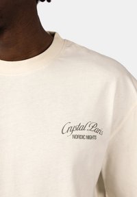Cream cotton long-sleeve shirt features a round neckline and printed text logo "Crystal Paris" with "NORDIC NIGHTS" in stylized gray lettering.