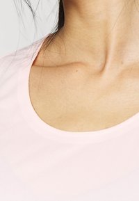 Primer plano del cuello y la parte superior del pecho de una mujer que lleva una camiseta de cuello redondo rosa claro, con mechones de cabello oscuro visibles.