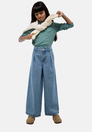 Pantalons en denim larges de couleur bleu clair avec une taille haute, associés à un haut à manches longues côtelé vert et une écharpe crème, portés de façon décontractée.