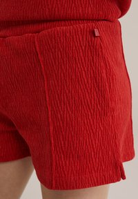Červené textúrované šortky s rebrovaným vzorom, s bočnou kapsou a malou logo etiketu na páske. Krátka dĺžka a uvoľnený strih.
