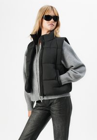 Vrouw in een zwarte puffervest, grijze trui, gestructureerde zwarte broek en zwarte zonnenbril, staand met één hand in de zak tegen een witte achtergrond.