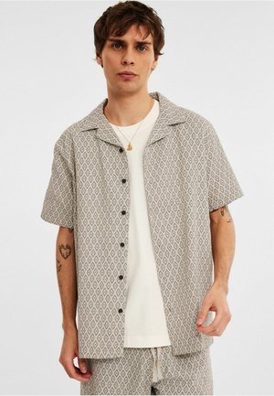 Koszula krótki rękaw, zapinana na guziki, w beżowym materiale z czarnym wzorem w romby; ma kołnierz typu spread i luźny krój. Nosi się ją na białym t-shircie.