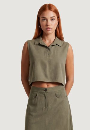 Femme aux longs cheveux roux porte un haut court sans manches vert olive et une jupe assortie, debout devant un fond clair uni.