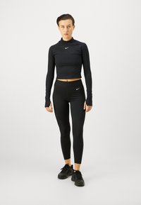 Schwarzes Sportset bestehend aus einem kurzärmligen Top mit hohem Kragen und schwarzen Leggings mit strukturierten Einsätzen, kombiniert mit schwarzen Sneakers.