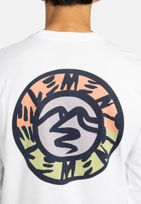 Camicia bianca a maniche lunghe con un grafiche circolare che presenta design astratti di montagne e mani in arancione, verde e nero. Materiale in cotone.