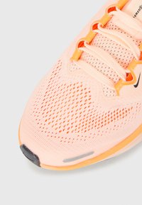 Chaussure de sport rose clair avec des accents orange, tige en mesh respirant, lacets et petit logo swoosh noir près des œillets de lacets.