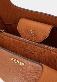 Braunes, strukturiertes Lederinnenfutter einer Guess-Handtasche mit Druckknopfbeutel, Reißverschlussfach und geprägtem Markenlogo innen und außen.
