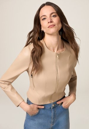 Giacca in ecopelle beige con scollo rotondo, chiusura con zip e tasche, abbinata a una gonna di jeans chiara. Tessuto liscio.
