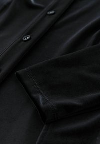 Schwarzes, weiches Stoffhemd mit knöpfbarem Vorderteil, glatter Textur und sichtbaren Nähdetails an den Ärmeln und dem Saum.