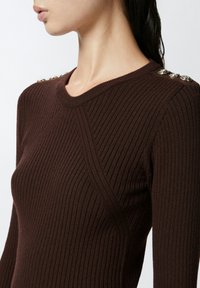 Maglietta marrone a maniche lunghe con scollo rotondo e dettagli in bottone metallico sulla spalla. Design aderente con tessuto testurizzato.