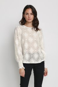 White lace blouse featuring intricate floral patterns, long sleeves with gathered cuffs, and a relaxed fit. Paired with black pants.