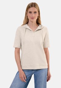 Ljusbeige hoodie med korta ärmar, framknäppning och dragsnören. Mjukt, texturerat tyg med avslappnad passform. Bärs tillsammans med blå jeans.