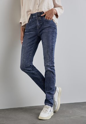 Donkerblauwe denim jeans met een slim fit, voorzien van twee voorzakken en een knoopsluiting, stijlvol gecombineerd met witte sneakers voor contrast.