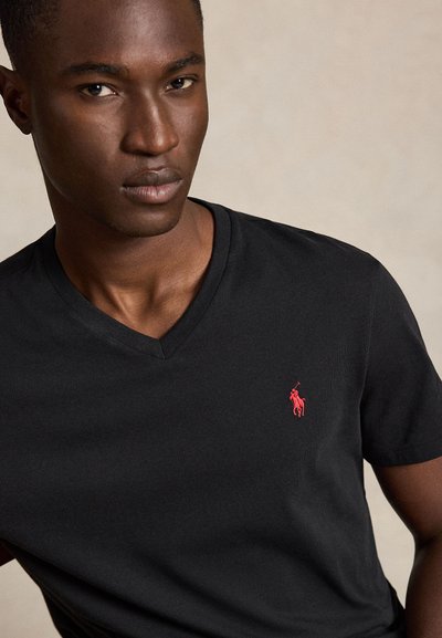 Mladi moški nosi črno majico z V-izrezom in majhno rdeče vezeno polo igralčevo značko na levi strani prsnega koša.