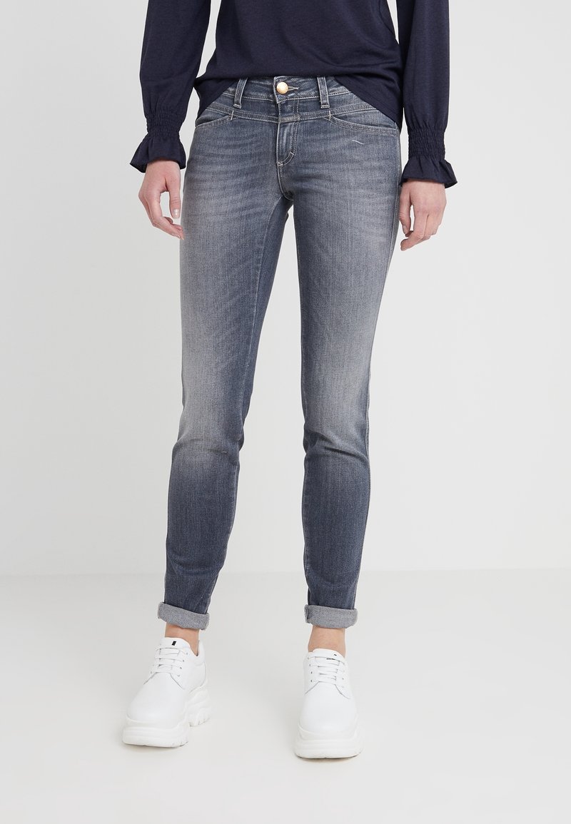 Personne portant un jean skinny gris retroussé, des baskets blanches et un haut manches longues bleu marine debout sur un fond clair uni.