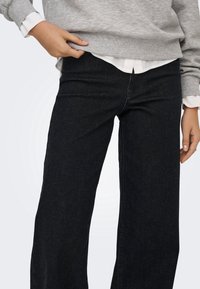 Personne portant un jean noir à jambes larges, un sweat gris et une chemise blanche, ajustant le sweat d'une main.
