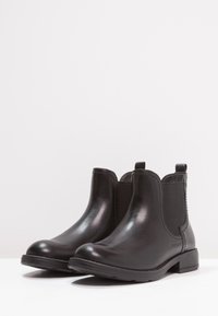 Un par de botas Chelsea de cuero negro con paneles elásticos laterales y tacones bajos sobre una superficie blanca y un fondo plano.