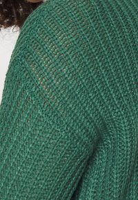 Grön stickad tröja med en texturerad, öppen struktur och diagonal ribbad design. Har ett mjukt, lätt tyg som är idealiskt för lager-på-lager.