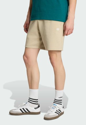 Person trägt beige Adidas-Shorts, weiße Adidas-Sneaker mit schwarzen Streifen, weiße Socken mit schwarzen Streifen und steht vor einfarbigem Hintergrund.