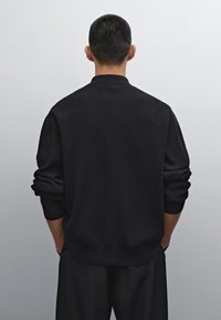 Schwarzer, gerippter Pullover mit entspanntem Schnitt, überschnittenen Schultern und langen Ärmeln, von hinten vor hellem Hintergrund zu sehen.