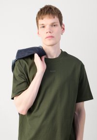 Olivgrünes Baumwoll-T-Shirt mit Rundhalsausschnitt, kurzen Ärmeln und dezentem "HUGO"-Logo, mit Denimstoff, der über die Schulter gelegt ist.