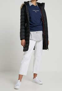 Abrigo largo negro acolchado con capucha de pelo sintético, sudadera azul marino con logo, camisa blanca con rayas debajo, vaqueros blancos recortados, zapatillas blancas.