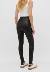 Leggings de cuero sintético negros con una cintura alta, textura suave y diseño elegante, combinados con una camiseta corta blanca y zapatillas casuales.