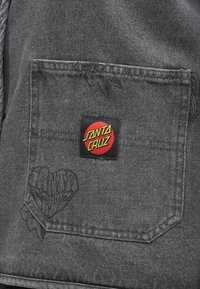 Svart denimjakke bak med et lite rødt og gult "Santa Cruz" logo-patch på den øvre lommen, med subtile blomsterdesign.