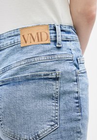 Εγγύς λήψη πίσω τσέπης φωτεινών μπλε τζιν με ταμπά δερμάτινη ετικέτα της μάρκας "VMD", που φοριέται με λευκό πουκάμισο.