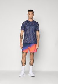 Navy-kuosisilla urheilupaidalla, jossa on lyhyet hihat, yhdistettynä sinisestä oranssiksi häivyttyviin shorts-eihin, ja varustettuna valkoisilla lenkkareilla.
