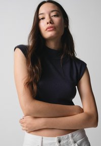 Young woman with long dark hair, wearing a sleeveless black top and white pants, arms crossed, looking confidently at the camera.