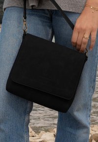 Schwarze Wildleder-Crossbody-Tasche mit Klappenverschluss, ausgestattet mit einem goldenen Hardware-Clip und detaillierten Nähten. Tragen mit hellblauen Jeans.