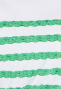 Witte stof met groene golfvormige borduursels in een herhalend patroon van golven, met complexe details en textuur.