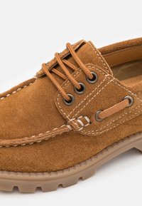 Suede ruskea vene-kengät, joissa on pyöreä kärki, kontrastivalkoiset tikkaukset ja ruskeat nauhat. Kumipohja, jossa on kuvioitu pito.