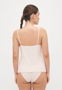 Lys rosa topp med tynne justerbare stropper og en bølget hem, laget av et glatt, mykt stoff. Matchende rosa truser er synlige under.