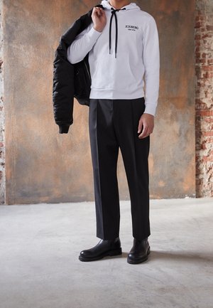Homme portant un sweat à capuche blanc avec le texte « ICEBERG », un pantalon de costume noir et des bottes noires, tenant une veste noire sur une épaule.