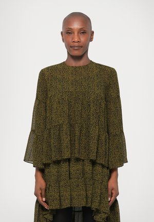 Robe superposée verte foncée avec un motif paisley jaune, manches longues évasées et silhouette fluide. Le tissu transparent ajoute de la texture.