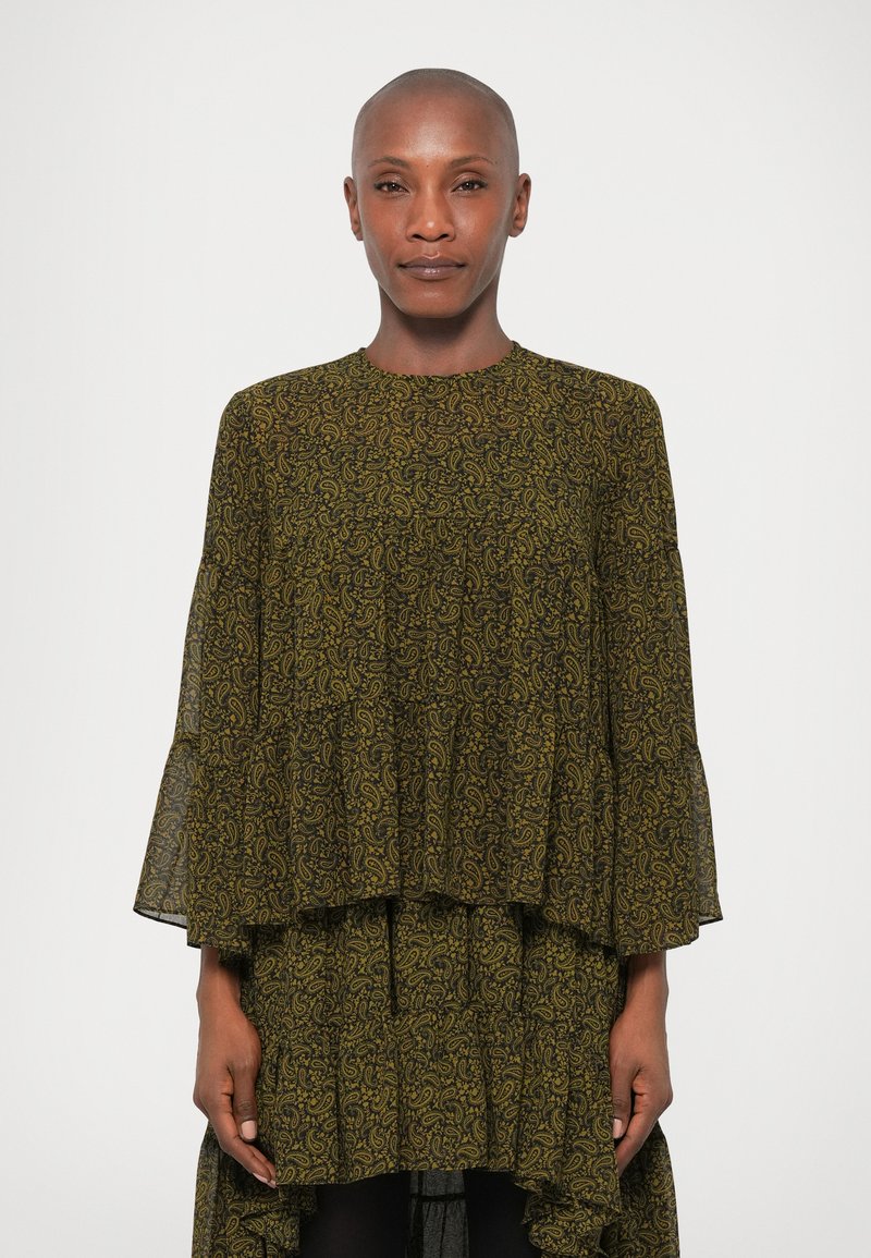 Dunkelgrünes, schichtweise gefertigtes Kleid mit gelbem Paisleymuster, langen, schwingenden Ärmeln und einer fließenden Silhouette. Der transparente Stoff sorgt für Textur.