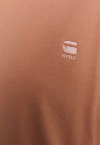 Ruskea puuvillapaita, jossa on ribbaustekstuuria ja pieni valkoinen brodeerattu logo vasemmalla puolella. Yksinkertainen, lyhythihainen muotoilu.