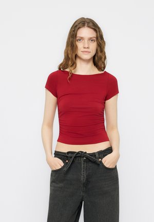 Ung kvinde med bølget hår har en tætsiddende rød kortærmet top og løse sorte jeans på med hænderne i lommerne, stående mod en hvid baggrund.