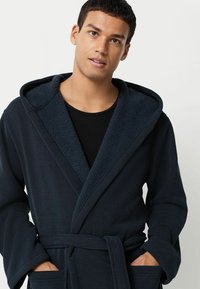 Tmavomodrý plášť so štruktúrovaným vonkajším materiálom a mäkkou, fleecovou kapucňou. Obsahuje pásový opasok a veľké vrecká.