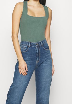 Femme portant un débardeur côtelé vert à larges bretelles et un jean bleu taille haute, debout devant un fond clair uni.