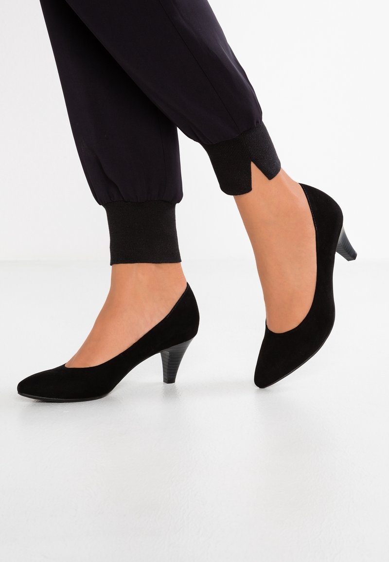 Escarpins en daim noir à talons moyens portés avec un pantalon noir retroussé sur pieds nus, sur un fond blanc uni.