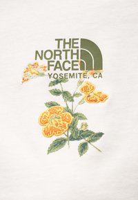Λευκό ύφασμα με το λογότυπο "The North Face" σε πράσινο κείμενο με κίτρινη κεντητική με λουλούδια, περιλαμβάνοντας πορτοκαλί λουλούδια και πράσινα φύλλα.