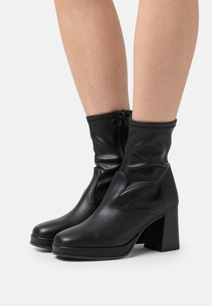 Bottes en cuir noires arrivant au mollet, à talons blocs et avec des fermetures éclair latérales, portées sur des jambes nues sur fond blanc.