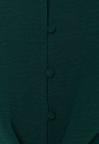 Tessuto verde scuro con una superficie morbida, caratterizzato da una fila verticale di quattro bottoni arrotondati abbinati e dettagli di cucitura visibili.