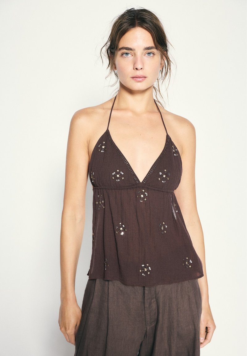 Jeune femme à la peau claire et aux yeux bleus portant un top dos nu marron avec des motifs floraux en sequins et un pantalon ample marron foncé.