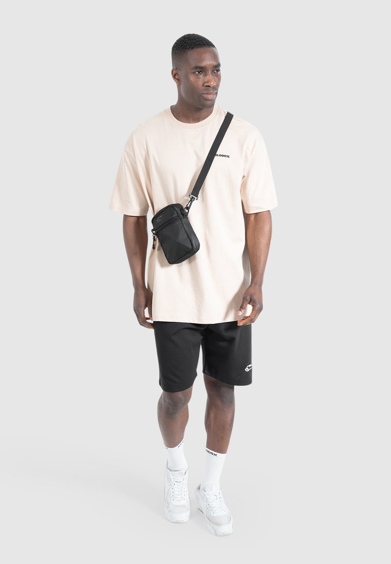 Black crossbody bag with two zip compartments, worn over a light pink oversized t-shirt and black shorts. White sneakers and socks accentuate the outfit.
